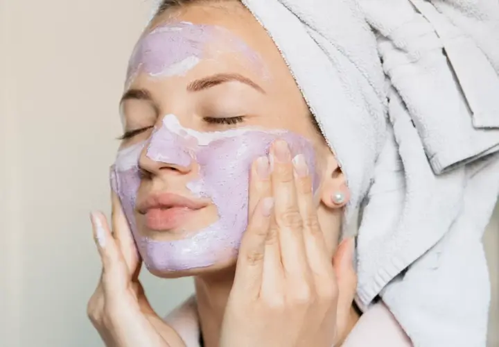 En kvinna med handduk i håret applicerar en lila ansiktsmask. Hon njuter av att ånga ansiktet för en avkopplande stund.