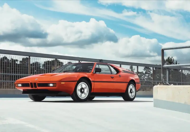 Pomarańczowe BMW E26, ikona lat 80., prezentuje się na tle błękitnego nieba z chmurami.