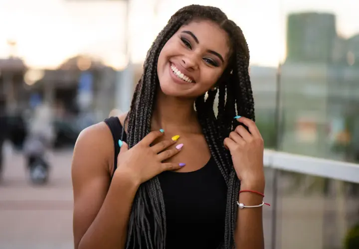 Joven sonriente con trenzas africanas, uñas de colores y un brazalete rojo.