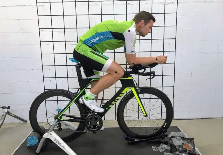 Mężczyzna w stroju kolarskim na rowerze stacjonarnym podczas sesji bike fitting. Punkty pomiarowe na ciele i rowerze.