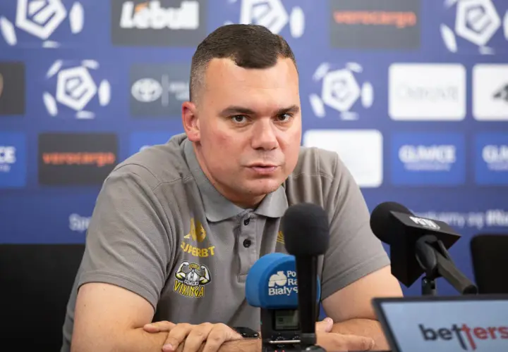 Trener Jagiellonii w szarej koszulce z logo klubu, siedzi przy mikrofonach podczas konferencji prasowej.