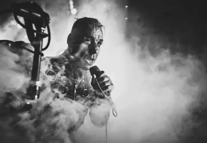 Lindemann w dymie, z mikrofonem w dłoni, w mrocznym klimacie. Jego twarz jest intensywna, jakby śpiewał "Knebel".