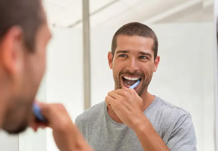 Uomo sorridente si lava i denti davanti allo specchio, un gesto quotidiano per la prevenzione odontoiatrica.