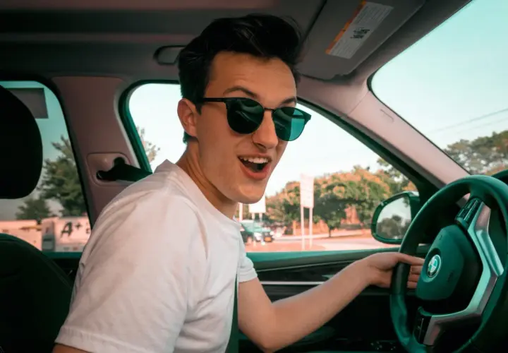 Joven con gafas de sol sonríe emocionado al volante de su coche novel, listo para una aventura.