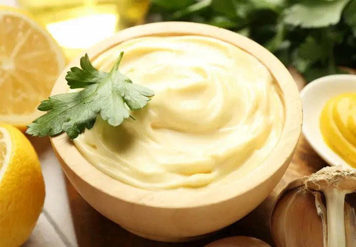 Mayonnaise onctueuse dans un bol en bois, garnie de persil. Citrons, ail et moutarde à proximité. La clé pour ne pas rater une mayonnaise maison.
