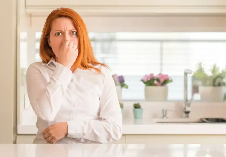 Mujer pelirroja se tapa la nariz, con cara de asco, por unos gases con olor a huevos podridos en la cocina.