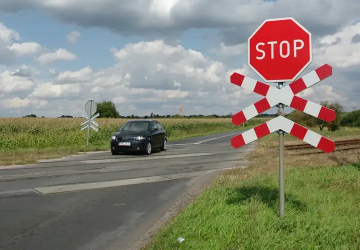 Czarny samochód zbliża się do przejazdu kolejowego z czerwonym znakiem STOP i białoczerwonym krzyżem.