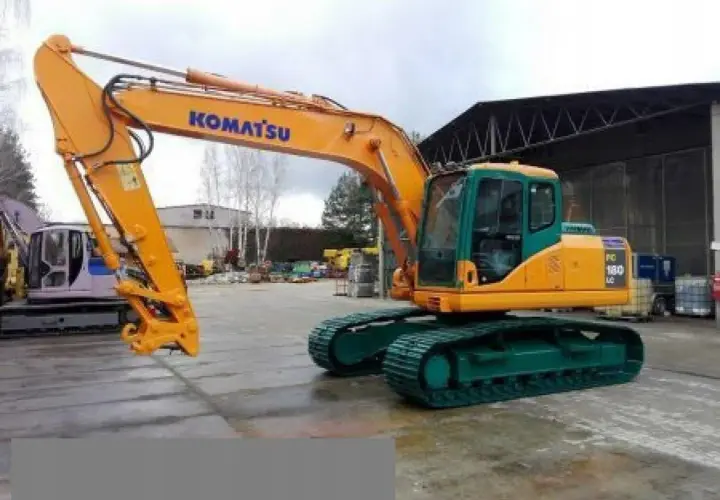 Pomarańczowa koparka gąsienicowa Komatsu PC 180 LC z zielonym podwoziem stoi na placu budowy.