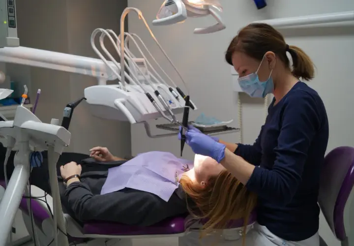 Dentystka w masce i rękawiczkach pracuje przy pacjencie. W gabinecie widoczny sprzęt do stomatologii.