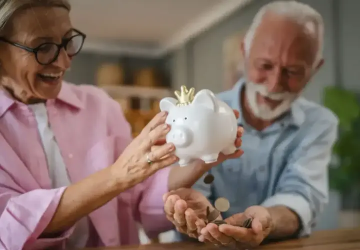 Uśmiechnięta para seniorów wsypuje monety do skarbonki-świnki. Czekają na moment, kiedy wyrównają emerytury czerwcowe.