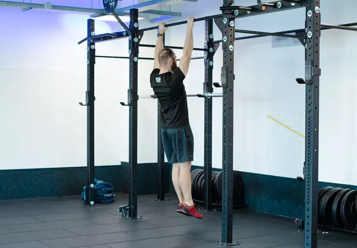Mężczyzna wykonuje scapular pull ups na drążku. Ćwiczy plecy i ramiona w siłowni.