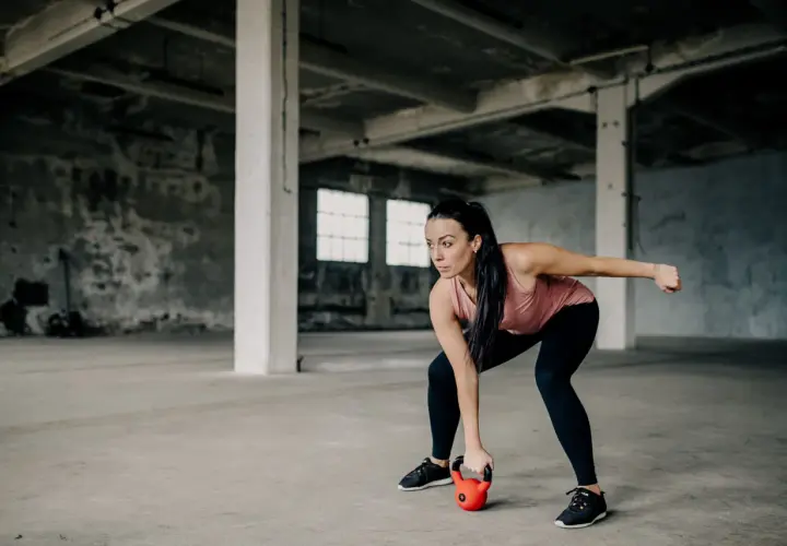 Kobieta w koszulce treningowej wykonuje ćwiczenie z kettlebell w opuszczonej hali.