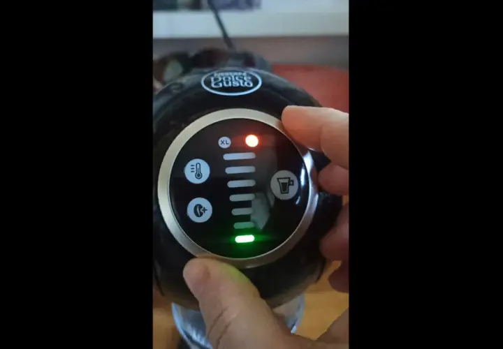 Dłoń naciska przycisk na ekspresie Dolce Gusto. Czerwona lampka sygnalizuje problem, a zielona wskazuje, że dolce gusto cieknie woda.
