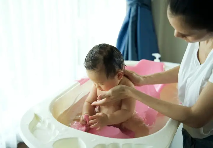 Mama delikatnie kąpie dziecko w wanience. Woda jest ciepła, a maluch bawi się zabawką. Może dodano krochmal do kąpieli dla miękkości skóry?