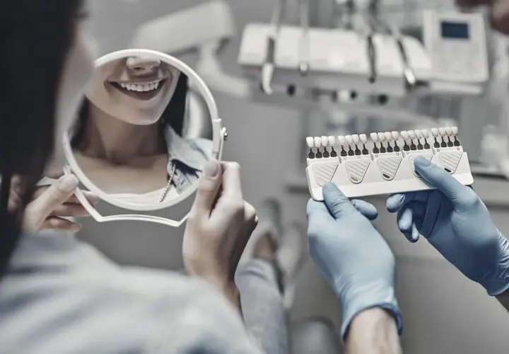 Dentysta w rękawiczkach pokazuje pacjentce wzornik kolorów zębów, pacjentka ogląda swój uśmiech w lustrze.