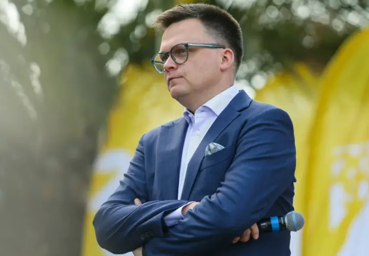 Szymon Hołownia w garniturze i okularach, z mikrofonem w ręku, na tle żółtych flag.