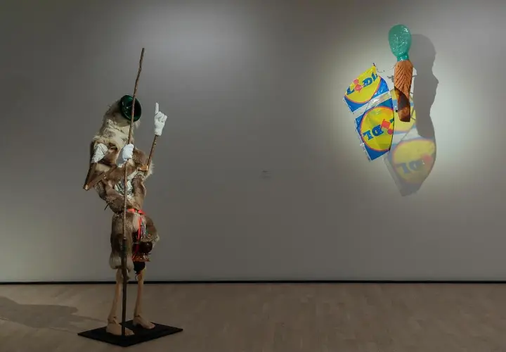 Konsthallar Sverige: En surrealistisk figur med päls och en pinne står bredvid en väggskulptur med Lidl-påsar och ett grönt huvud.