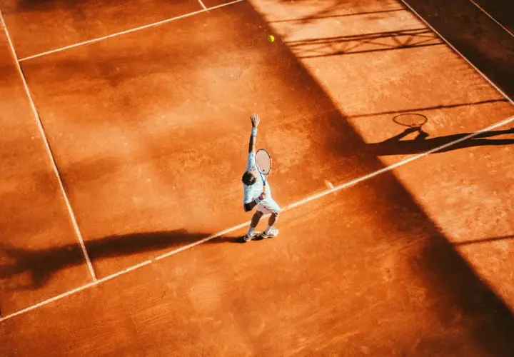 Agassi tenisista w akcji, serwuje na ceglanym korcie. Cień rakiety i sylwetka gracza rzucają się na pomarańczową nawierzchnię.