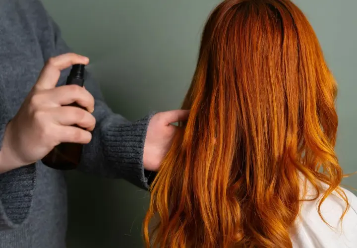 Persona rocía un producto en el cabello fino y quebradizo de color naranja rojizo para darle brillo y protección.