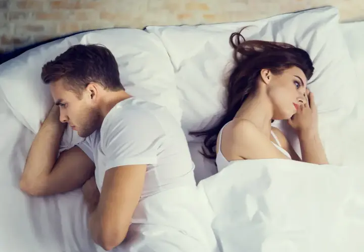 Coppia a letto, schiena contro schiena, pensierosi. A volte si può stare insieme senza fare l'amore, ma la distanza emotiva è palpabile.