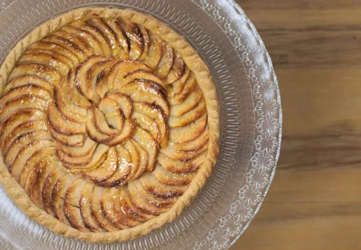 Un magnifique appareil tarte aux pommes, aux pommes caramélisées disposées en spirale sur une pâte dorée, sur un plat en verre décoré.
