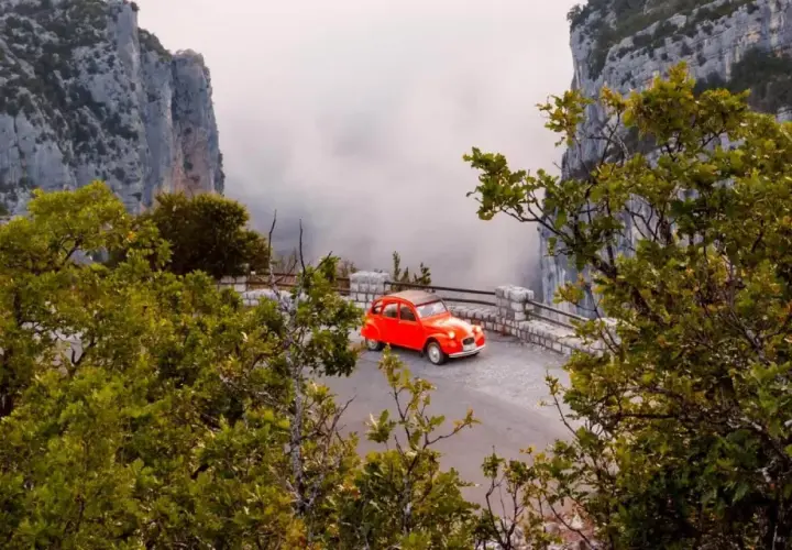 Une petite voiture rouge se gare sur la route des Gorges du Verdon, offrant une vue spectaculaire sur les falaises et la brume.