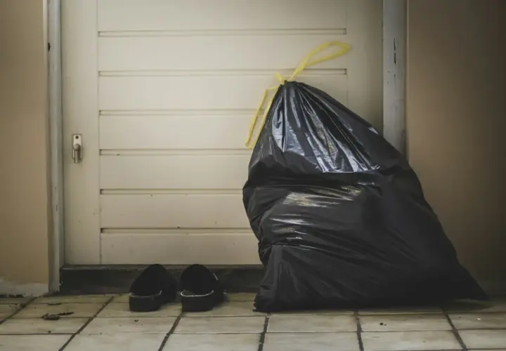 A full black trash bag and a pair of slippers sit by a closed door, symbolizing the need for lean budgeting to discard unnecessary expenses.