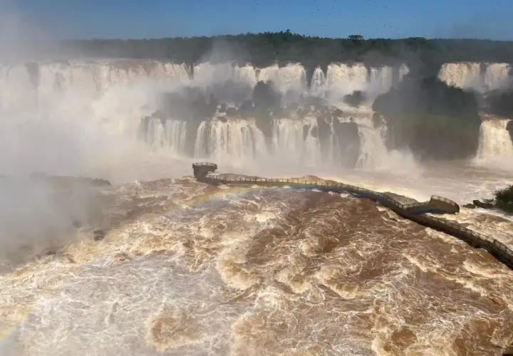 Mächtige Iguazú-Wasserfälle stürzen in schlammbraunes Wasser. Ein Steg führt durch den Sprühnebel, ein Regenbogen funkelt.