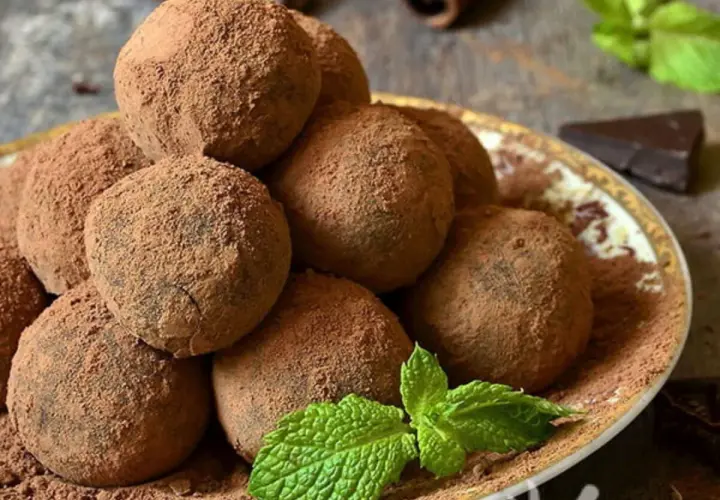 Un mucchio di tartufi al cioccolato facili e veloci, spolverati di cacao, su un piatto decorato, con menta fresca e un pezzetto di cioccolato fondente.