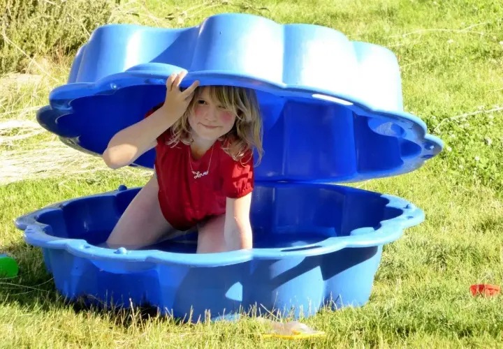 Niña sonríe bajo una tapa azul de arenero, disfrutando de actividades de verano. ¡Diversión acuática para niños de 2 años!