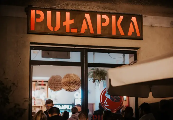 Neonowy napis "PUŁAPKA" nad wejściem do lokalu. W środku widać ludzi i dekoracje.