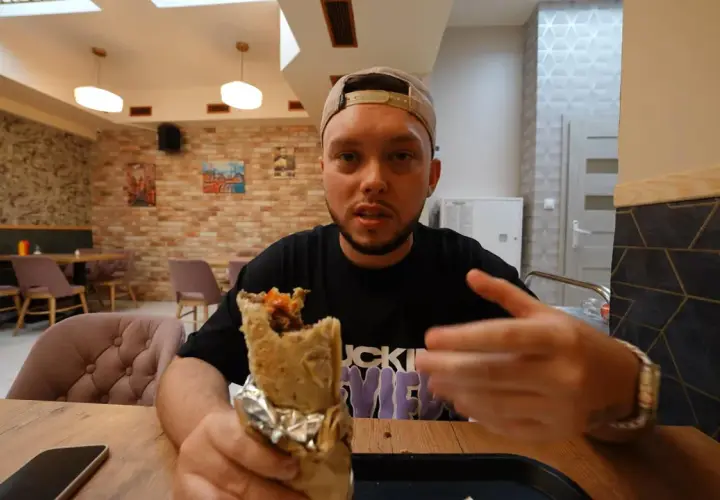 Mężczyzna z kebabem w ręku, prawdopodobnie najlepszy kebab warszawa. Wnętrze restauracji z ceglanymi ścianami i wygodnymi fotelami.
