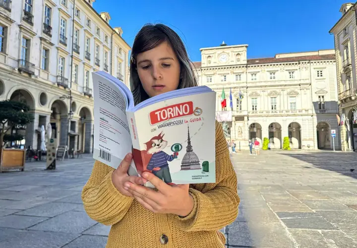 Bambina legge una guida "Torino con Viaggiapiccoli" in una piazza torinese. La libreria viaggi è perfetta per scoprire la città.