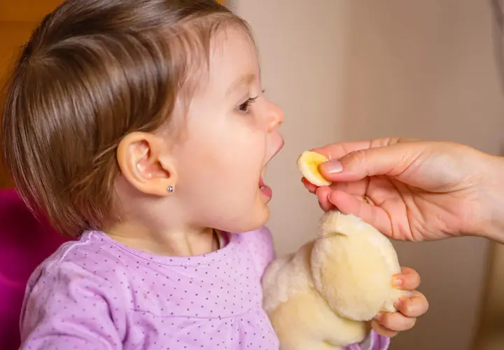 Małe dziecko z otwartymi ustami czeka na kawałek banana. Po biegunce u dziecka ważne jest nawadnianie i lekkie posiłki.