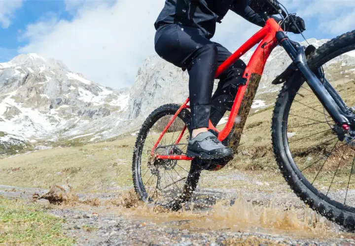 Kolarz na czerwonym rowerze trekkingowym pokonuje błotnistą kałużę na górskim szlaku.