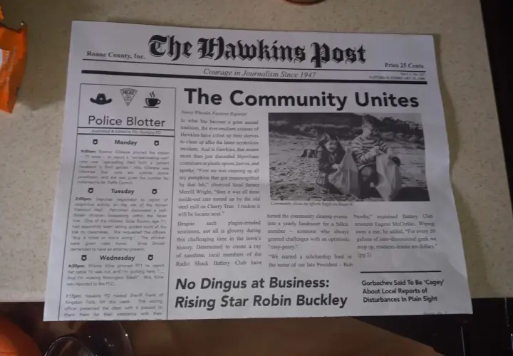 Gazeta "The Hawkins Post" z artykułem "The Community Unites" i zdjęciem dzieci sprzątających plażę.