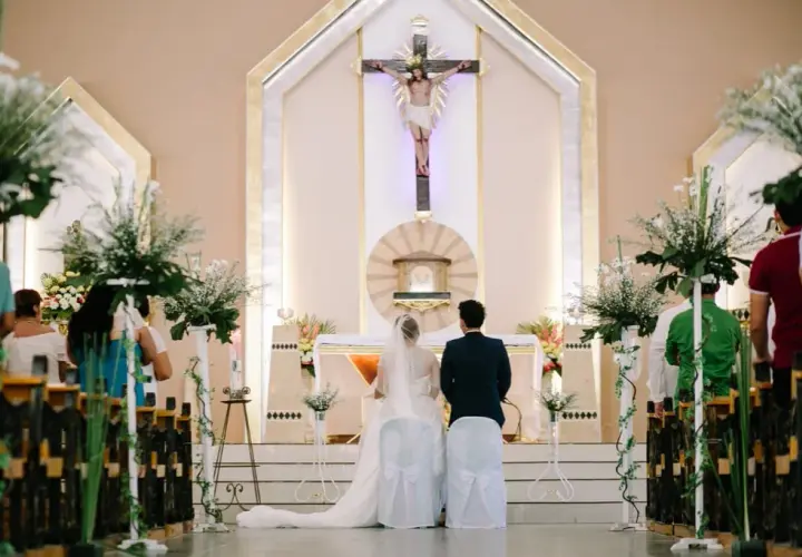 Le déroulement du mariage à l'église commence avec le couple devant l'autel, sous un crucifix. Les invités sont assis dans les bancs décorés de fleurs.