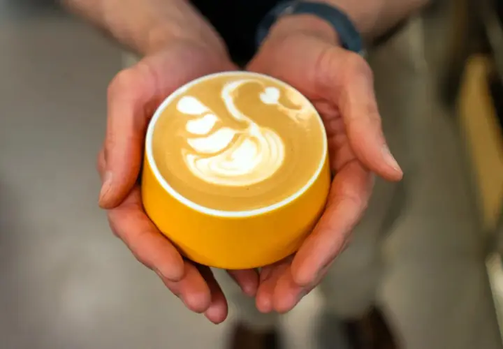 Dłonie trzymają żółtą filiżankę z kawą, na której artystycznie uformowano łabędzia. Zastanawiasz się, ile kofeiny ma espresso z ekspresu?