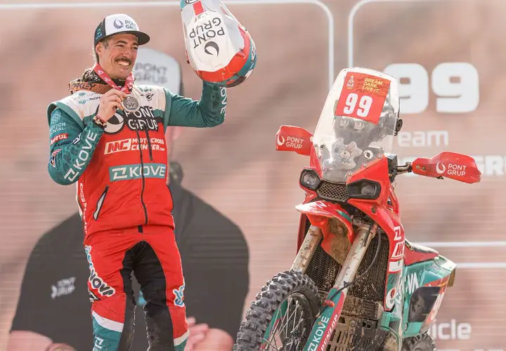 Piloto celebra victoria junto a su moto china Dakar. Casco y medalla en mano, luce orgulloso su equipación.