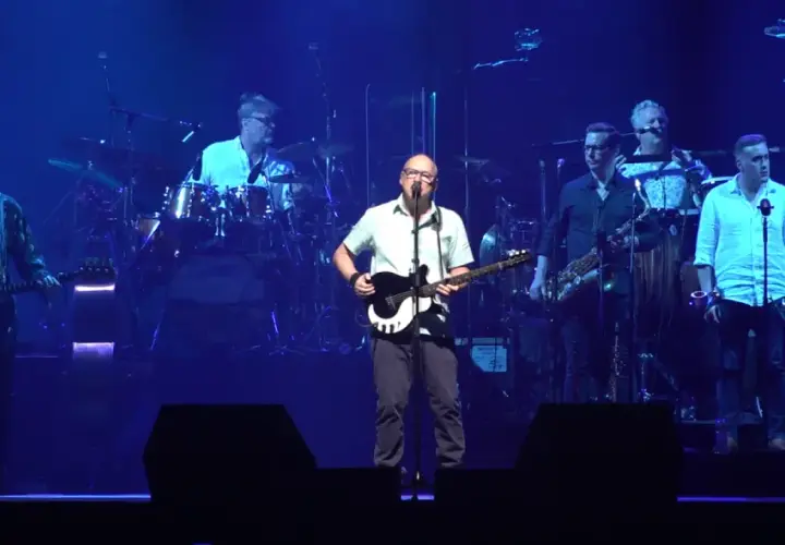 Mark Knopfler na scenie z zespołem podczas jednego z jego niezapomnianych koncertów. Muzycy grają na gitarach, saksofonach i perkusji.