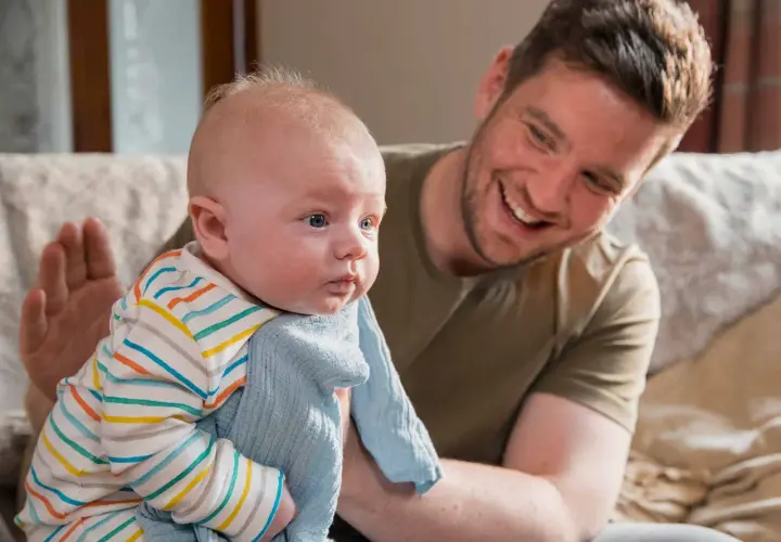 Padre sorridente tiene in braccio un neonato con una copertina azzurra. La posizione del neonato sembra confortevole, forse per alleviare il singhiozzo.