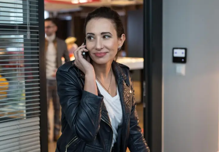 Aleksandra Popławska w skórzanej kurtce rozmawia przez telefon. W tle widać mężczyznę i biurowe wnętrze, przypominające scenę z jej filmów i seriali.