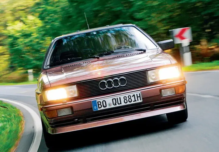 Klasyczny, brązowy Audi quattro z lat 80. pędzi po krętej, leśnej drodze.