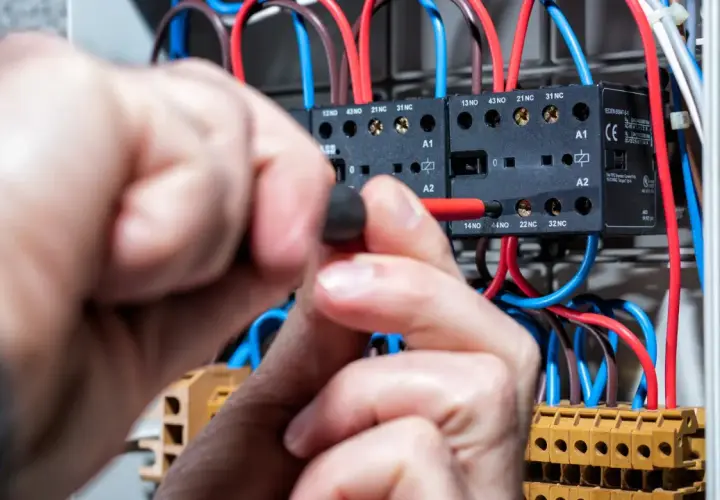 Ręce elektryka pracują przy rozdzielnicy, podłączając przewody. Widać styki i bezpieczniki, a napięcie w sieci jest pod kontrolą.