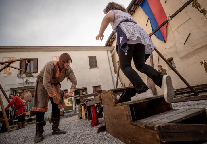 Bambini si divertono con giochi medievali. Un uomo in costume d'epoca aiuta una bambina a salire su una struttura di legno.