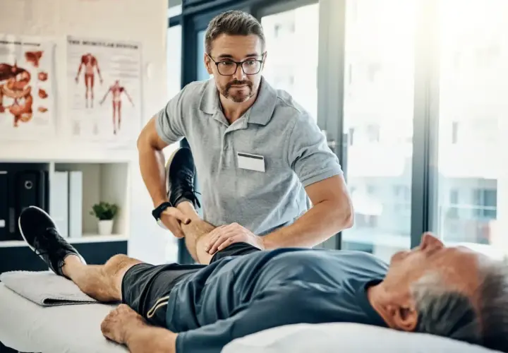 Fizjoterapeuta wykonuje świadczenie rehabilitacyjne, pomagając starszemu pacjentowi w ćwiczeniach nogi.