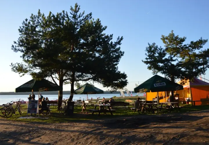 Relaks na plaży Karolinów. Ludzie siedzą pod parasolami, rowery czekają na plaży, a w tle jezioro i namioty.