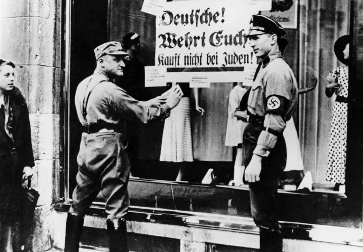 Ein SA-Mann klebt ein Plakat "Deutsche! Wehrt Euch! Kauft nicht bei Juden!" an ein Schaufenster. Dies ist ein Beispiel für den **Alltag im Nationalsozialismus**.