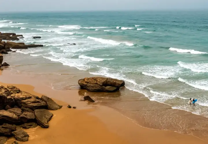 Maroko plaże: skaliste wybrzeże, piaszczysta plaża i turkusowe fale Atlantyku. Dwóch surferów wchodzi do wody z deskami.