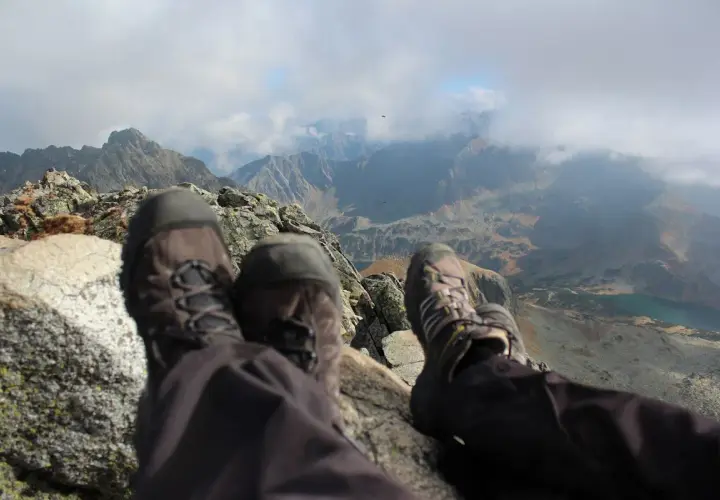 Stopy w butach trekkingowych na tle jesiennych Tatr. Październikowe słońce przebija się przez chmury, oświetlając skaliste szczyty i turkusowe jezioro.